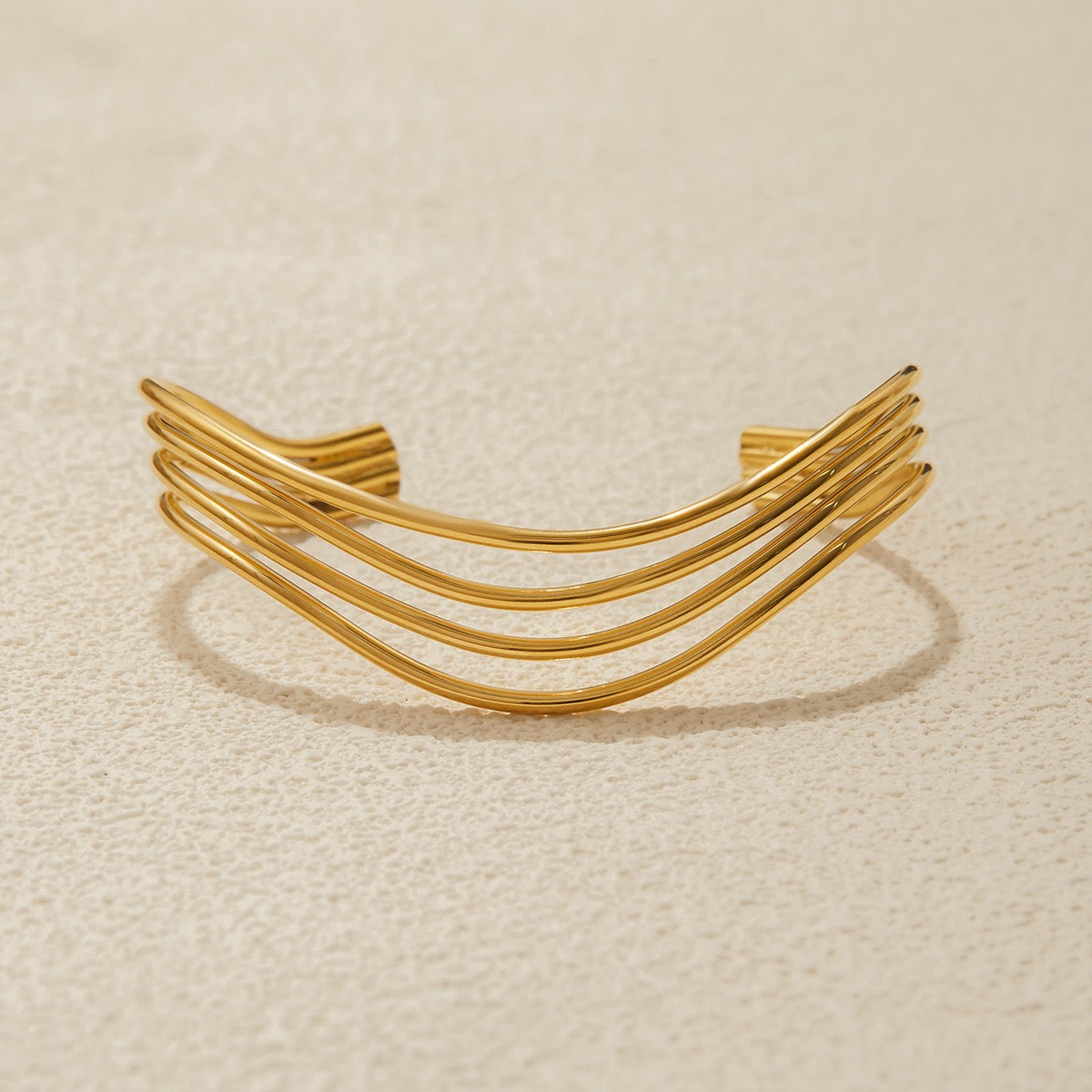 A gold-toned open cuff bracelet with three parallel curved bands, photographed on a textured light beige surface.